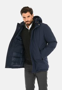 Giacca puffer blu navy con cappuccio, dotata di chiusura a zip e tasche laterali, foderata internamente con texture trapuntata e dal taglio morbido.