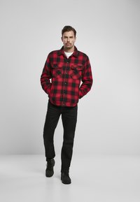 Chaqueta de camisa a cuadros rojos y negros con dos bolsillos en el pecho, cierre de botones y mangas largas, combinada con jeans negros y botas.