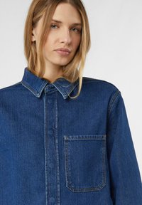 Koszula jeansowa w intensywnym niebieskim kolorze z zapięciem na guziki, ostrym kołnierzykiem i pojedynczą kieszenią na piersi. Charakteryzuje się kontrastowym przeszyciem i luźnym krojem.