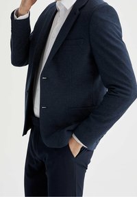 Blazer strutturato blu navy con revers a punta, due tasche frontali e due bottoni tono argento. Abbinato a una camicia bianca e pantaloni scuri.
