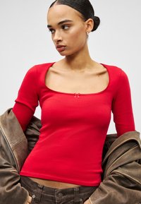 Crveni top s dugim rukavima i četvrtastim izrezom, s malim mašnom u sredini, u kombinaciji s smeđom jaknom. Glatka, uska tekstura.