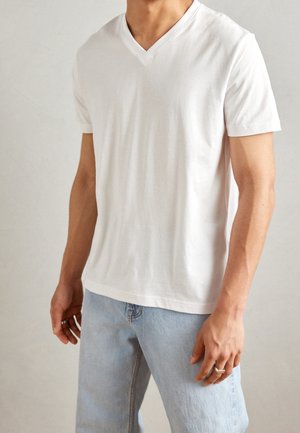 Homme portant un t-shirt blanc uni à col en V et un jean bleu clair, debout devant un fond neutre, les mains détendues le long du corps.
