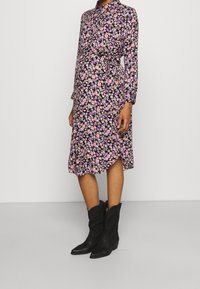 Květinové šaty s černým pozadím, zdobené růžovými a fialovými květinami, s dlouhými rukávy, límečkem na knoflíky a páskem v pase. Nosené s černými cowboyskými botami.