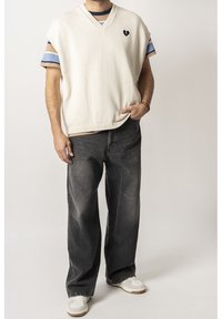 Cremesfarbener ärmelloser Strickpullover mit V-Ausschnitt, versehen mit einem kleinen schwarzen Herz-Logo, kombiniert mit lockeren, ausgewaschenen schwarzen Jeans und weißen Sneakers.