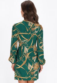 Zöld ruha aranylánccal és virágmintával, hosszú ujjakkal, begyűrt mandzsettákkal és testhezálló sziluettel, hátul cipzárral.