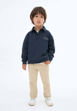 Pull assorti marine avec un col à boutons et un logo, accompagné de pantalons beiges et de baskets blanches. Tissu lisse avec une coupe décontractée.