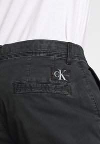 Černé bavlněné kalhoty s hladkou texturou, s zadní kapsou a značkovou nášivkou s bílým nápisem "Calvin Klein Jeans".