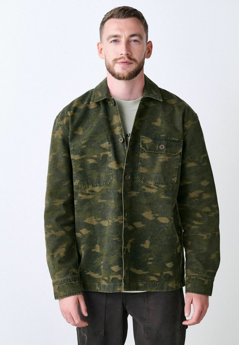Veste à motif camouflage vert, fabriquée en coton, avec un devant boutonné, deux poches poitrine et une coupe décontractée.