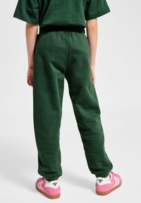 Grüne Baumwoll-Sweatpants mit lockerem Schnitt, ausgestattet mit einem elastischen Bund und schmal zulaufenden Beinen. Kombiniert mit rosa Turnschuhen.