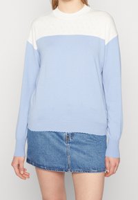 Ljusblå tröja med en vit, texturerad yoke. Långa ärmar, scallop-formad halsringning och en nederkant som sitter vid midjan. Matchas med en denimkjol.