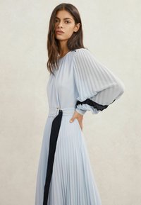 Rochie plisată bleu deschis cu mâneci lungi și dungi. Dispune de o curea în talie cu un accent negru. Textură netedă, design elegant.