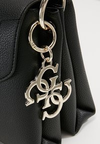 Mustan tekstuurin nahkaista käsilaukkua, jossa on kultainen avaimenperä, jossa on toisiinsa linkitettyjä kirjaimia. Rakenne on jämäkkä ja siinä on taitettavalla läpällä varustettu malli.