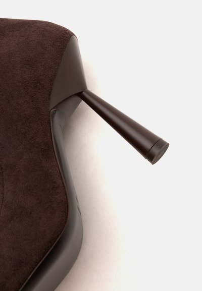Gros plan d'une chaussure à talon haut en daim marron montrant le talon aiguille fin et la semelle extérieure incurvée.