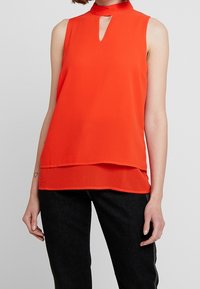 Červená bezrukávová blůza s vysokým límcem, s vykrojením ve tvaru klíče a vrstveným designem. Hladká tkanina a volný střih vytvářejí plynulou siluetu.
