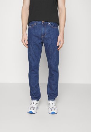 Mann trägt blaue Jeans mit geradem Bein, weiße Sneaker mit blauen Akzenten und ein schwarzes T-Shirt, steht vor einem schlichten Hintergrund.