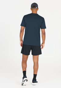 Man med svart keps, marinblå kortärmad skjorta, svarta shorts, svarta strumpor och sneakers som går bortåt på vit bakgrund.