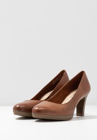 Scarpe con tacco alto in pelle marrone, con punta arrotondata, caratterizzate da una superficie liscia e un tacco in legno. Ambientazione interna su uno sfondo chiaro.