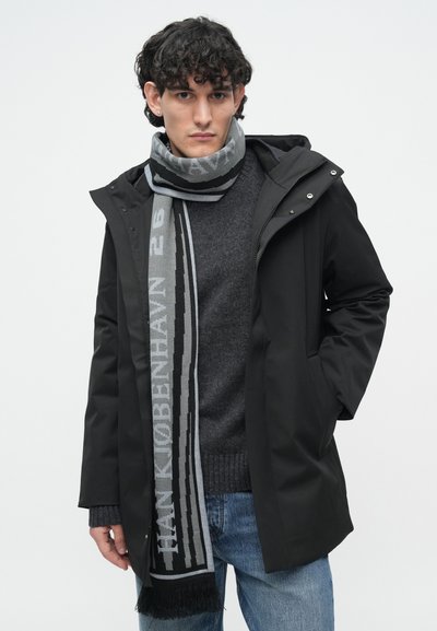 Jachetă neagră cu glugă, pulover tricotat gri dedesubt și o eșarfă gri și neagră cu text și detalii cu franjuri. Blugi casual din denim.