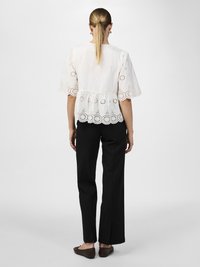 Blusa bianca a peplo con rifiniture ricamate, orlo a scalloped e maniche a sbuffo; abbinata a pantaloni neri a gamba dritta e ballerine marroni.