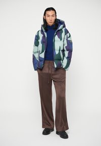 Homem de jaqueta puffer camuflada em azul-esverdeado sobre um suéter de gola alta navy, calças largas castanhas e ténis pretos, em pé contra um fundo branco.