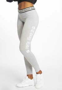 Grijze leggings met een zwarte "Thug Life" tailleband en witte grafische tekst op het linkerbeen, gecombineerd met witte sneakers. Glad materiaal.