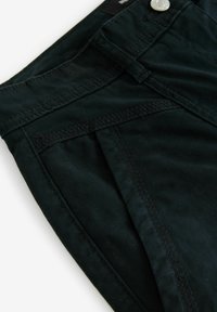 Pantalons en tissu vert foncé avec des coutures symétriques, des poches avant et un bouton en métal. La texture semble lisse et légèrement mate.