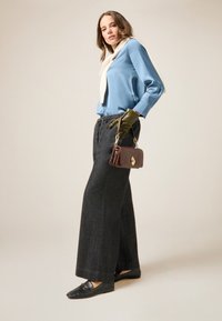 Himmelblaue Satinbluse, schwarze weite Denim-Hosen, grüne Lederhandschuhe und eine braune Handtasche mit goldenen Beschlägen. Neutraler Hintergrund.