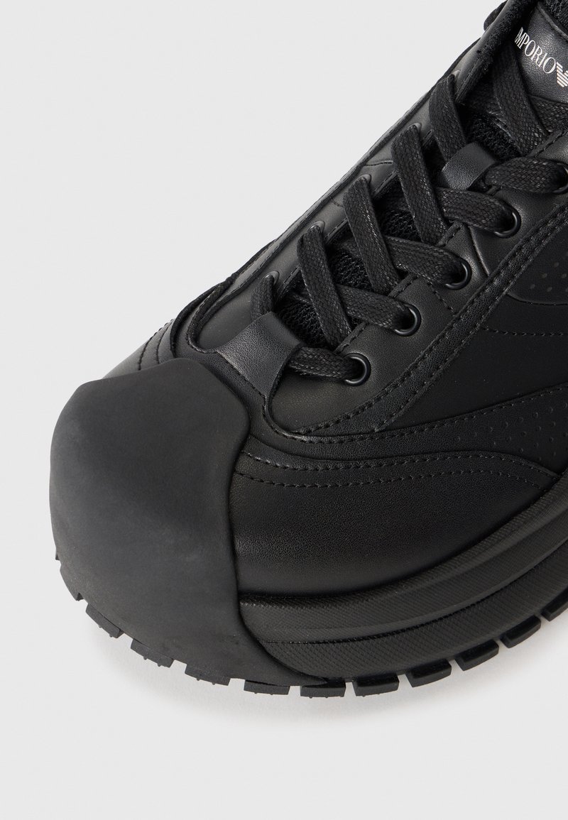 Svart idrottssko med en strukturerad läderöverdel, förstärkt gummitå, snörningsdesign och kontrasterande paneldetaljer.