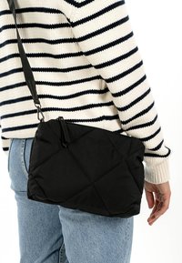 Person wearing blue jeans and white-and-black striped sweater carrying a black quilted crossbody bag with zipper closure.