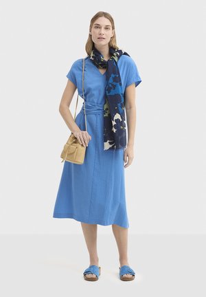 Robe midi bleue à côtes avec manches courtes, taille cintrée, associée à une écharpe florale foncée, un sac bandoulière beige et des sandales bleues.
