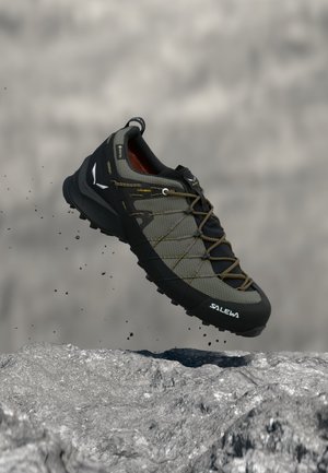 Chaussure de randonnée Salewa en olive et noir en plein vol au-dessus d'un terrain rocheux, montrant la semelle texturée et la tige lacée en matériau respirant.