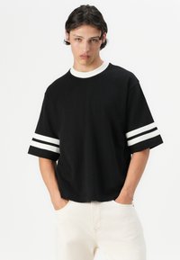Sort bomulds T-shirt med korte ærmer, der har hvide dobbelte striber på ærmerne og en kontrasterende hvid krave. Casual pasform.