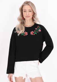 Felpa nera con ricami floreali in rosa e verde nella parte anteriore, dotata di maniche lunghe e colletto girocollo, abbinata a pantaloncini bianchi.