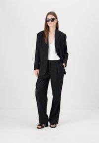 Svart blazer med en enkel knapp, kombinert med høyhårede svarte bukser og en hvit dyp V-hals topp. Fullført med svarte solbriller og sandaler.