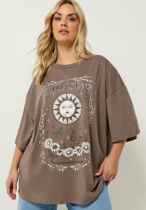 Kobieta w za dużym brązowym t-shircie z twarzą słońca, motylami, kwiatami i tekstem "SŁOŃCE" oraz "Twoje istnienie to magia."