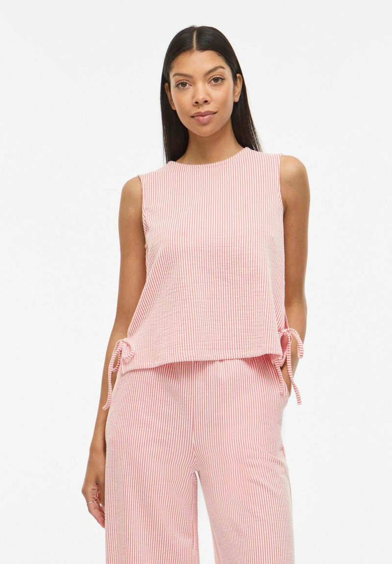 Femme aux longs cheveux foncés portant un haut sans manches rayé rose et blanc et un pantalon assorti, debout devant un fond blanc.