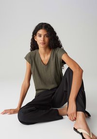 Olive green pleated top with a V-neck and short sleeves, paired with loose black pants and flat sandals featuring a beige strap.