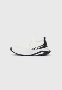Zapatilla deportiva blanca con una parte superior de tejido texturizado, detalles en negro, suela gruesa y un logotipo negro prominente en el lateral.