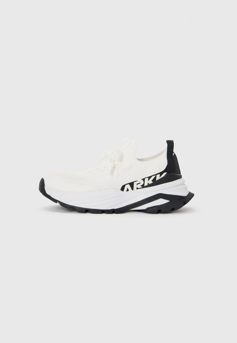 Zapatilla deportiva blanca con una parte superior de tejido texturizado, detalles en negro, suela gruesa y un logotipo negro prominente en el lateral.