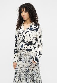 Πουκάμισο με κουμπιά σε κρεμ απόχρωση με μοτίβα που μοιάζουν με μαύρο μελάνι, μακριά μανίκια και άνετη εφαρμογή, σε συνδυασμό με φούστα με παρόμοιο μοτίβο.