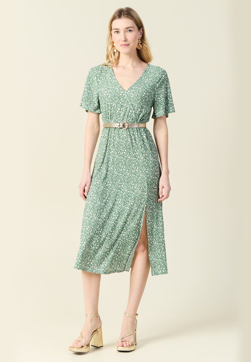 Zielona kobieca sukienka w kwiaty z krótkimi bufkami, dekoltem w serek, średniej długości i z bocznym rozcięciem, przewiązana w talii srebrnym paskiem.