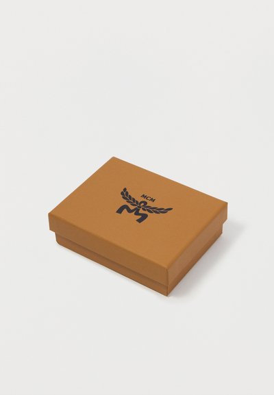 Καφέ χαρτοκιβώτιο με καπάκι, που φέρει ένα μαύρο λογότυπο με φτερά και "MCM" στην κορυφή. Τετράγωνο σχήμα, απαλός χειρισμός.