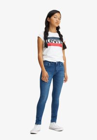 Camiseta blanca con un logo rojo y azul marino, jeans ajustados azules y zapatillas blancas. El atuendo presenta un diseño casual y entallado con una textura suave.