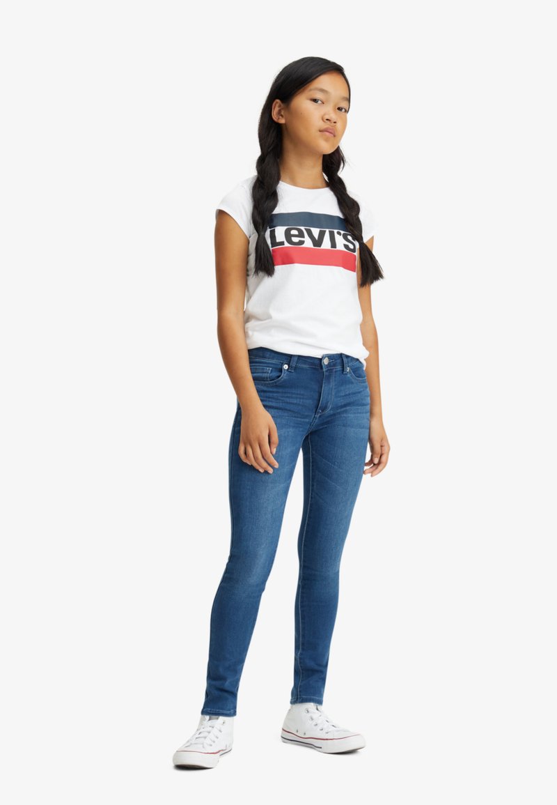 Camiseta blanca con un logo rojo y azul marino, jeans ajustados azules y zapatillas blancas. El atuendo presenta un diseño casual y entallado con una textura suave.