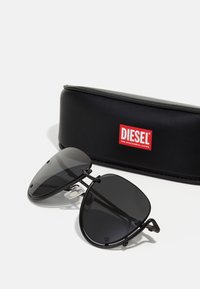 Lunettes de soleil aviateur noires avec verres sombres, monture en métal et coussinets de nez. Comprend un étui rigide noir avec un logo "DIESEL" rouge.