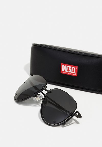 Lunettes de soleil aviateur noires avec verres sombres, monture en métal et coussinets de nez. Comprend un étui rigide noir avec un logo "DIESEL" rouge.