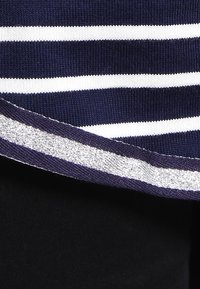 Marinblå randig stickad topp med vita horisontella ränder och en silverfärgad strukturerad accent längs nederkanten. Materialet verkar mjukt med en lätt lyster.