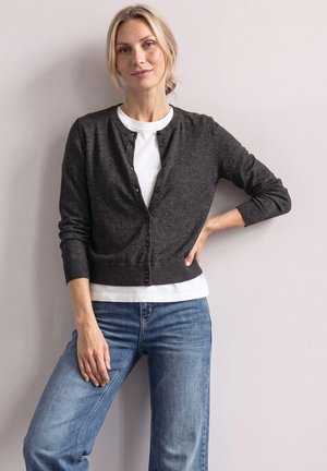Femme blonde debout contre un mur, portant un cardigan gris foncé boutonné sur une chemise blanche et un jean bleu, une main sur la hanche.