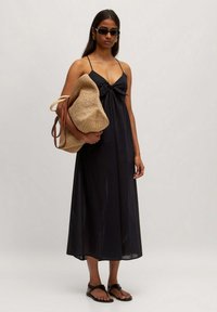 Schwarzes Maxikleid mit Knotendetail an der Brust, dünnen Trägern und fließender Silhouette. Dazu eine geflochtene Strohtasche und flache Sandalen.