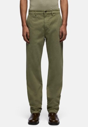 Man draagt olijfgroene rechte broek en donkerbruine leren schoenen, staand tegen een eenvoudige lichte achtergrond.